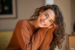 Joyful woman with long brown curly hair smiling and resting her head on her hands