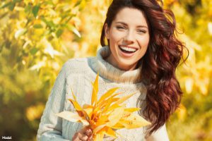 Woman with long red hair smiling outdoors while holding a bundle of fall leaves