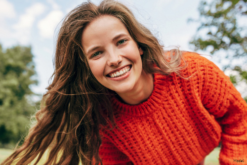 Smiling woman with long brown hair wearing an orange sweater outdoors