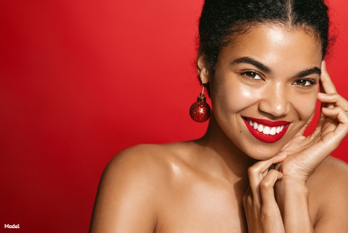Woman wearing red lipstick and red ornament earrings smiling and gently touching her face