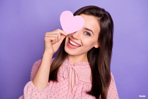 Joyful woman in a pink shirt holding a pink paper heart in front of her eye