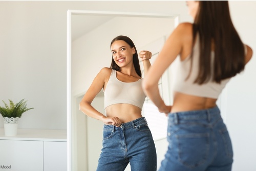 Woman in a sports bra and jeans smiling and pointing at herself in the mirror