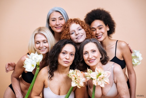 Six women of different ethnicities and ages posing together and smiling while holding white flowers